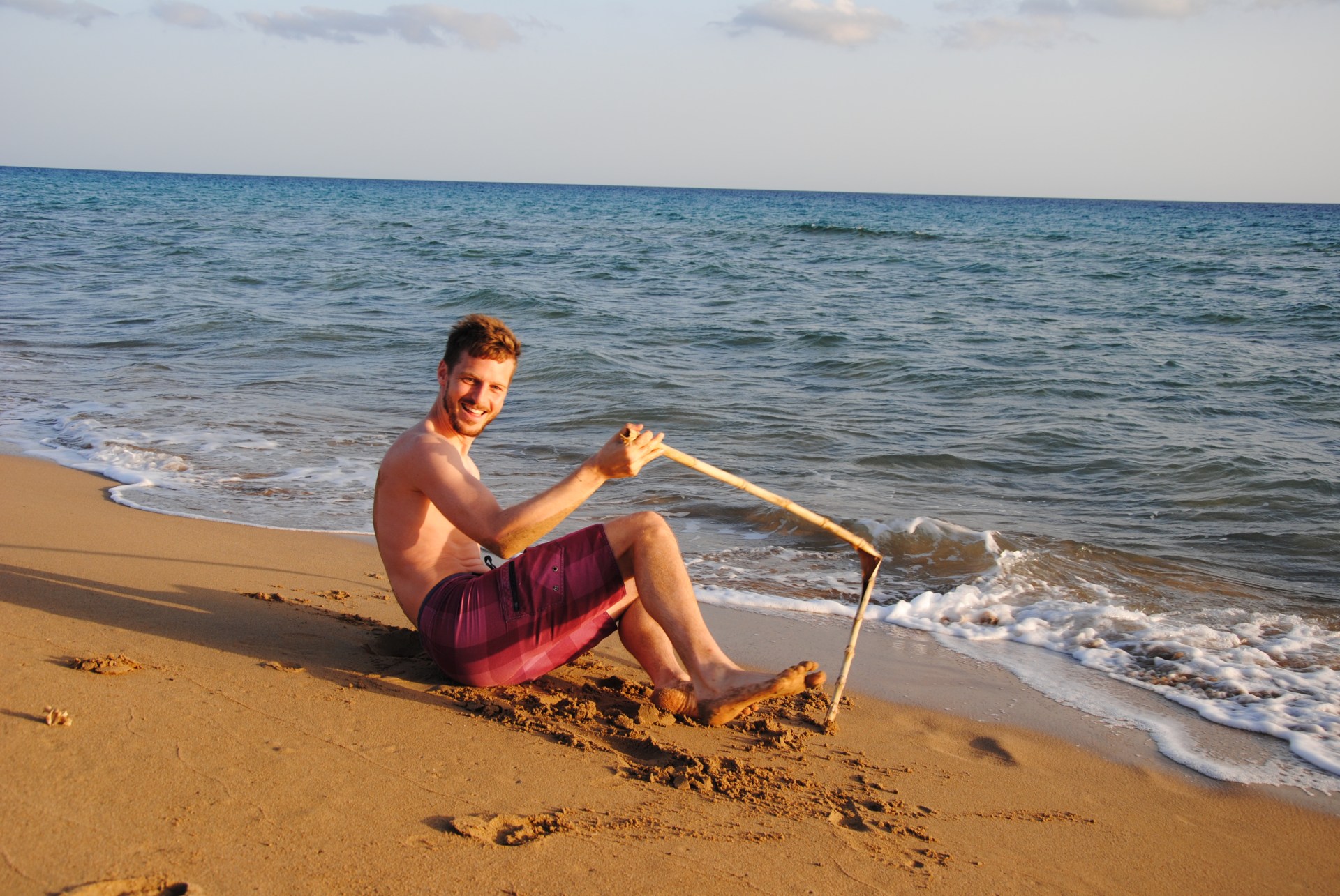 Sebastian liegt lachend am Strand und hält einen kaputten Stock