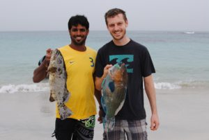 Sebastian steht mit einem Fischer am Meeresufer. Beide halten je einen Fisch in der Hand.