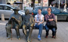 Leo und Hristina sitzen neben zwei Statuen auf einer Bank