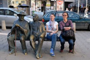 Leo und Hristina sitzen neben zwei Statuen auf einer Bank