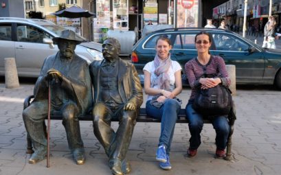 Leo und Hristina sitzen neben zwei Statuen auf einer Bank