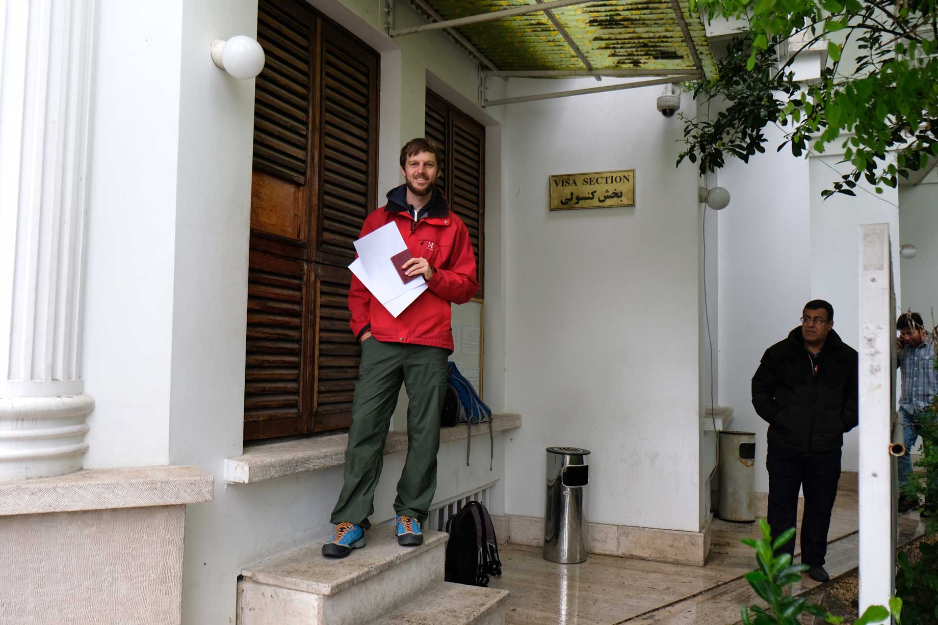 Sebastian steht mit Pass und Papieren in der Hand vor einem weißen Gebäude mit einem Schild, auf dem "Visa section" steht