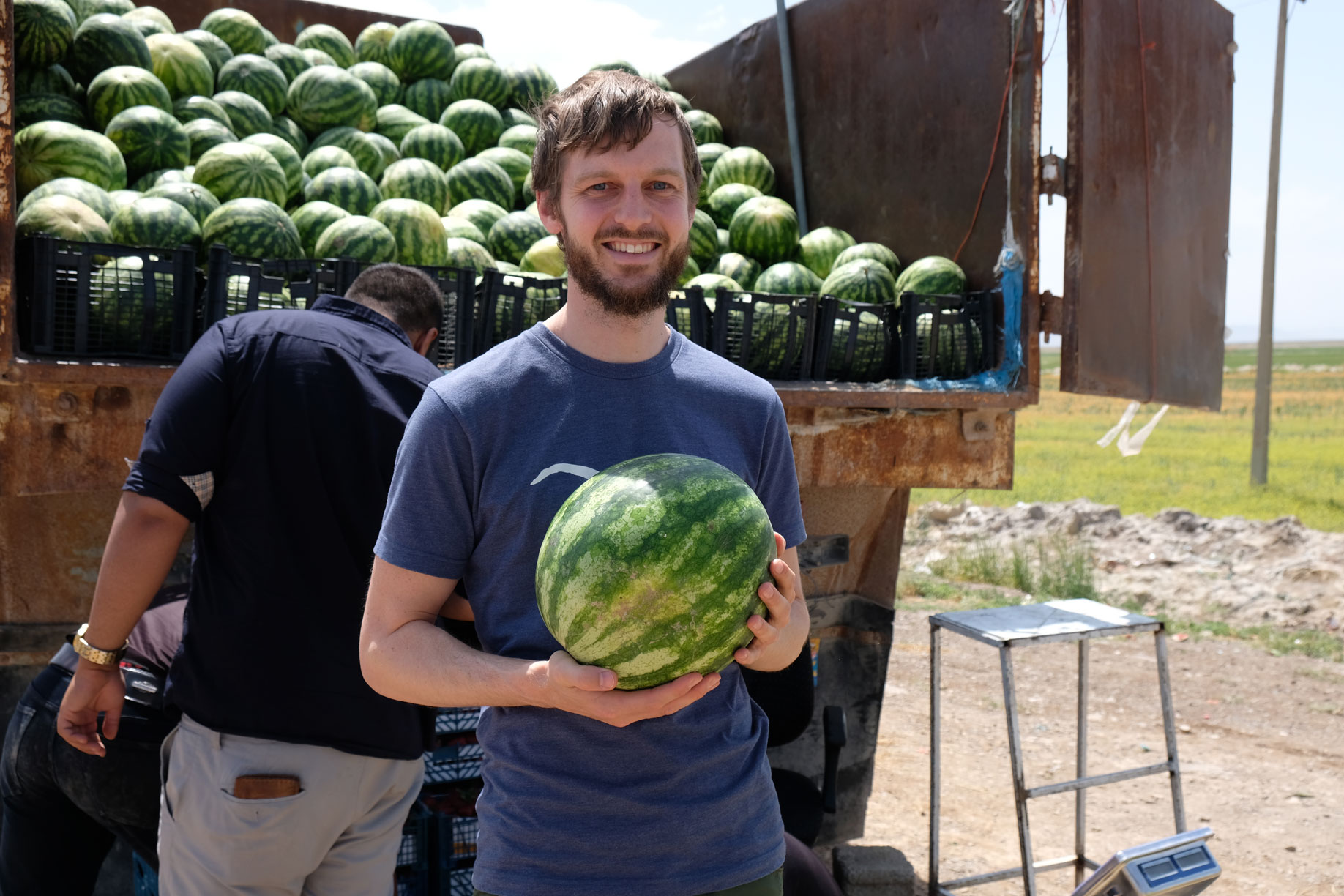 Sebastian hält vor einem Lastwagen, der mit Wassermelonen beladen ist, eine Wassermelone in der Hand