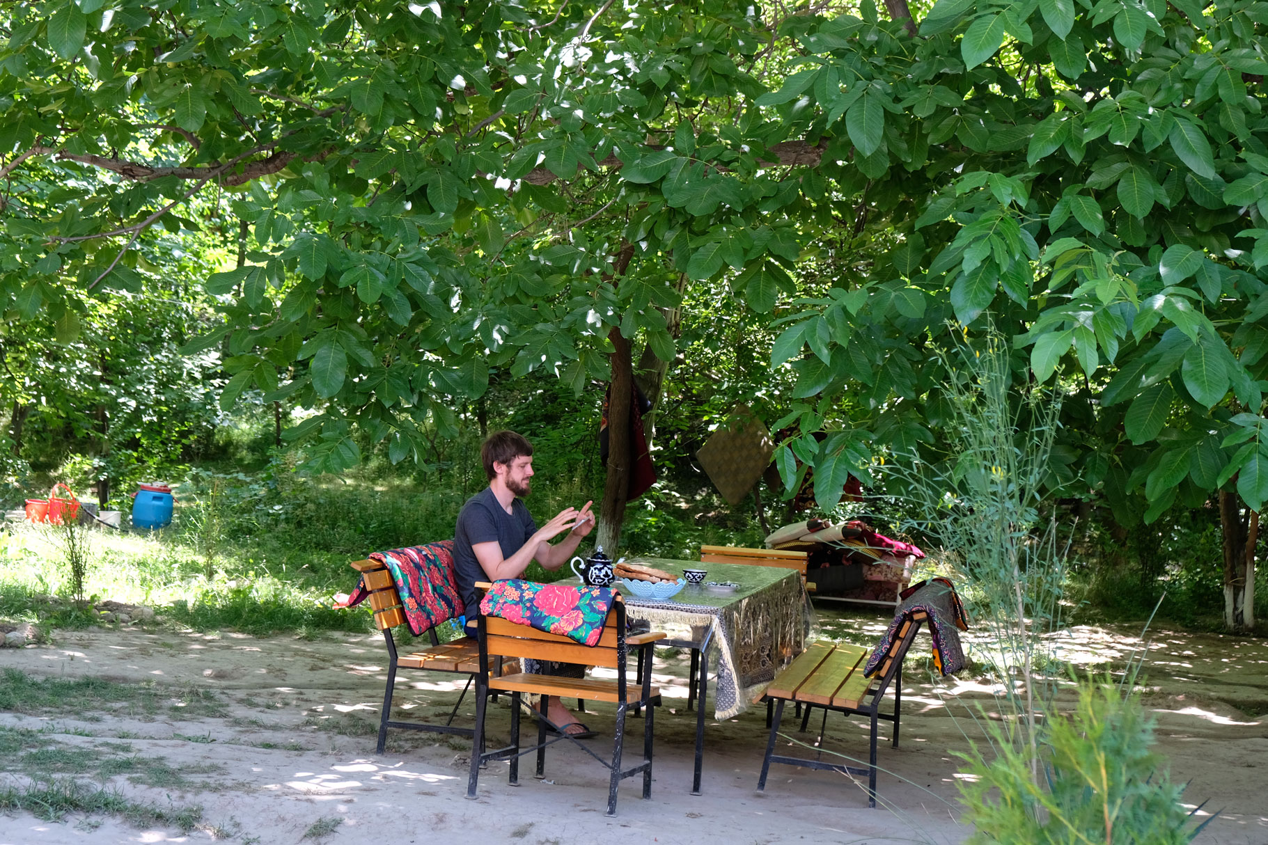 Sebastian sitzt mit Handy in der Hand an einem Tisch, der unter einem Baum steht.