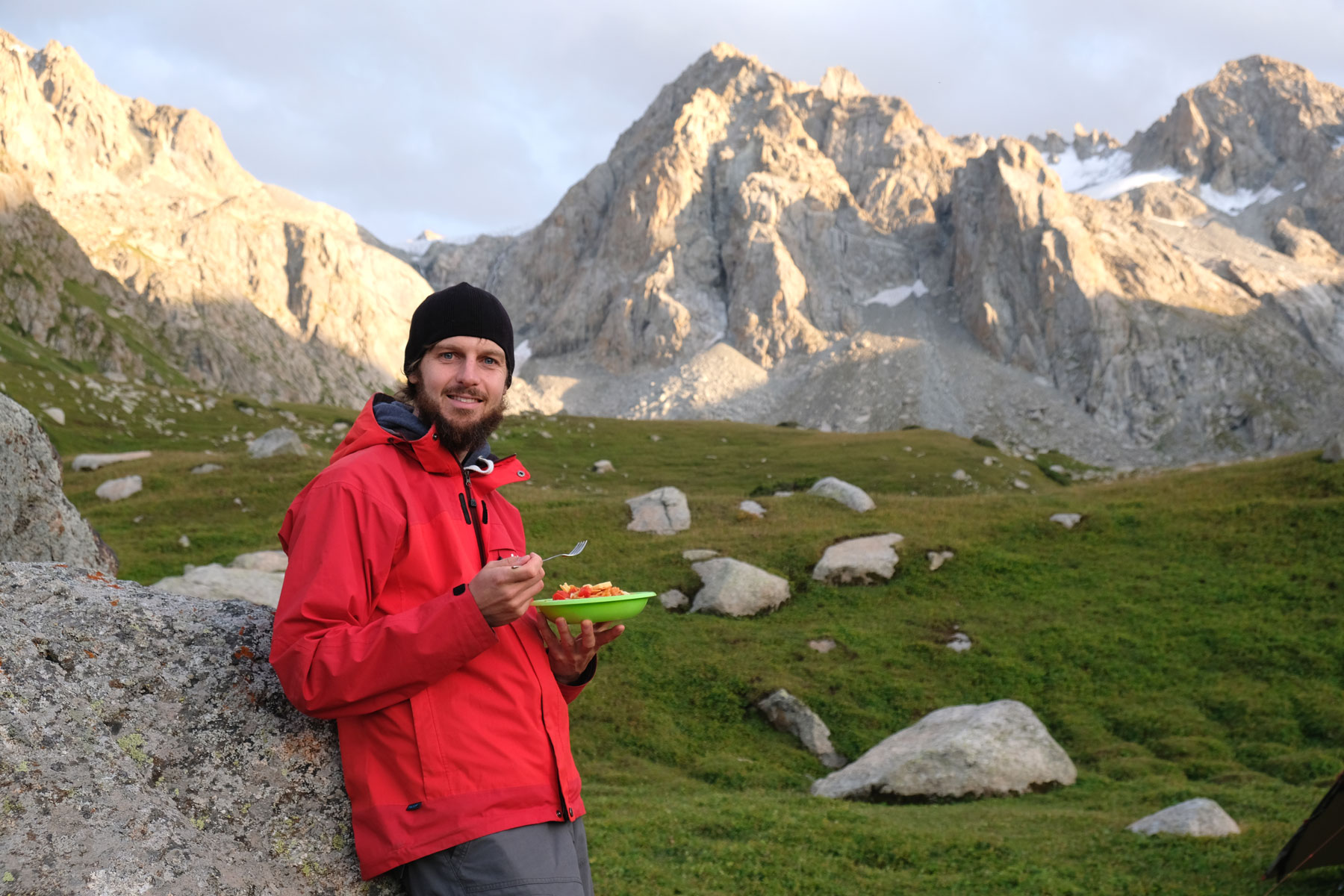 Sebastian leht neben an einem Felsen und in einer Gebirgslandschaft und hält einen Teller mit Essen in der Hand.