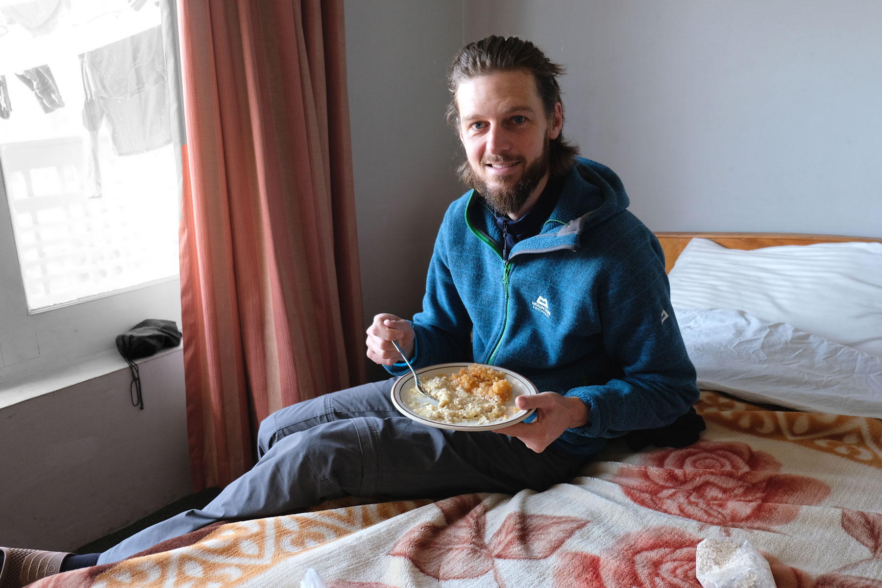 Sebastian sitzt auf einem Bett und hält einen Teller mit einem Brei aus Äpfeln und Bananen in der Hand.