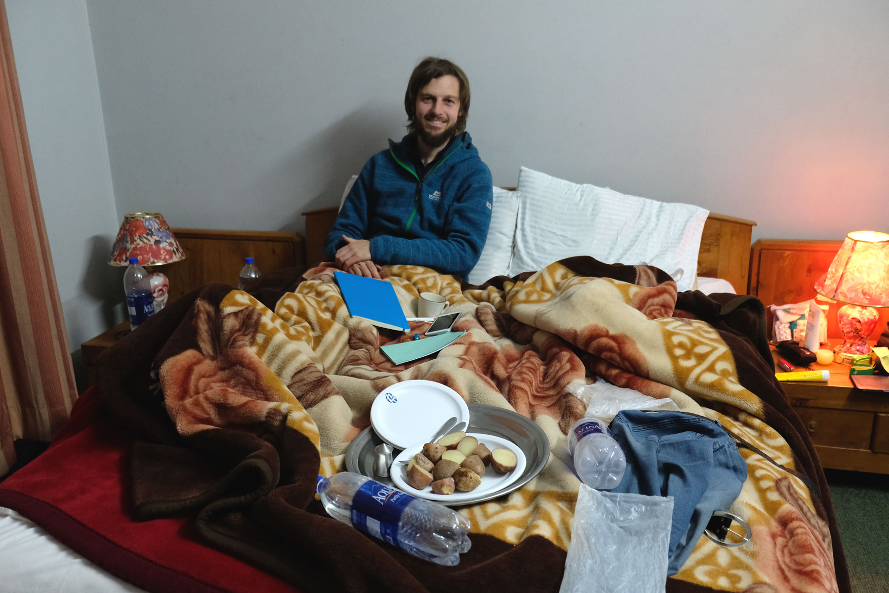 Sebastian sitzt in einem Bett, auf dem Wasserflaschen, ein Buch und ein Teller Kartoffeln liegen.