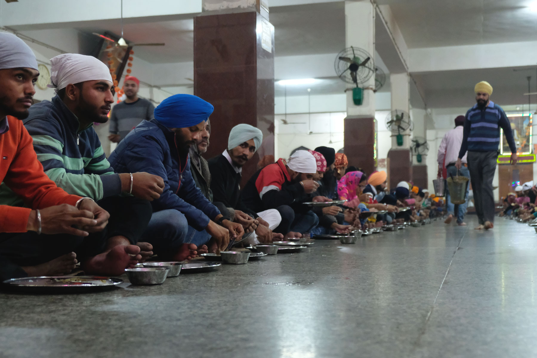 Menschen sitzen im goldenen Sikh-Tempel in Amritsar auf dem Boden. Vor ihnen steht Essgeschirr aus Metall.