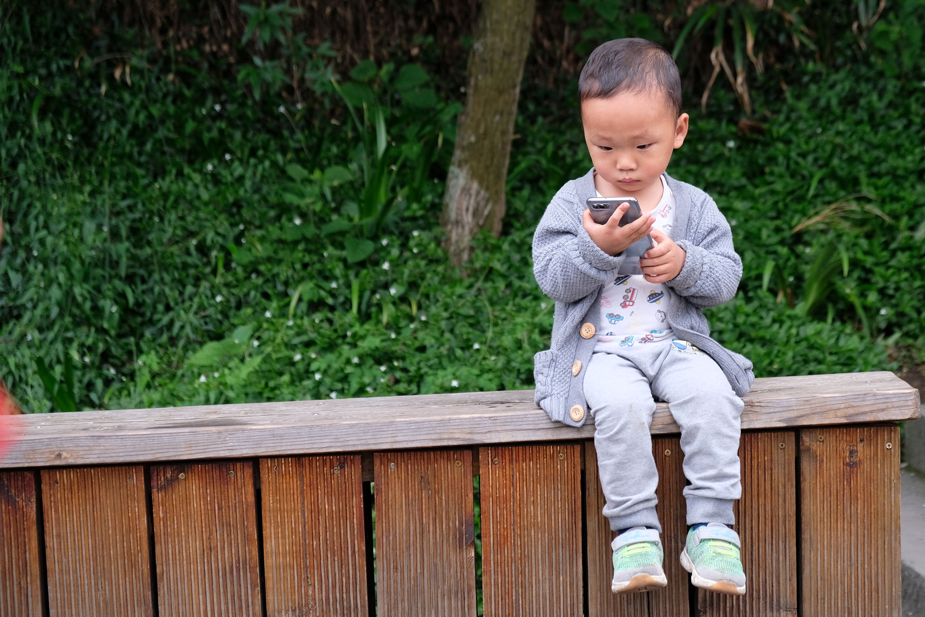 Ein chinesischer Junge sitzt auf einer Holzbank und hält ein Handy in der Hand.