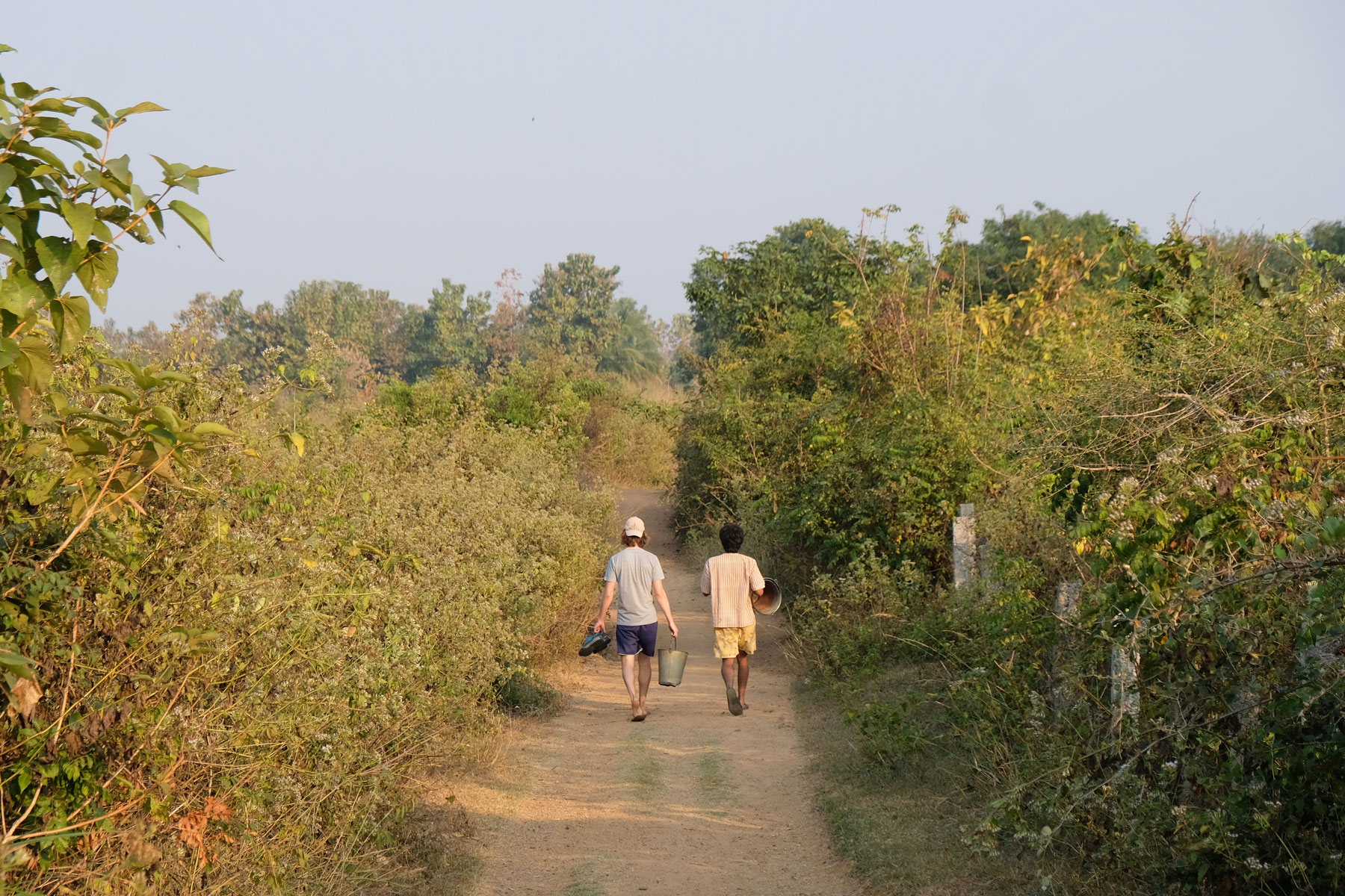Sebastian und Krish gehen mit Eimern in der Hand auf einem Feldweg.