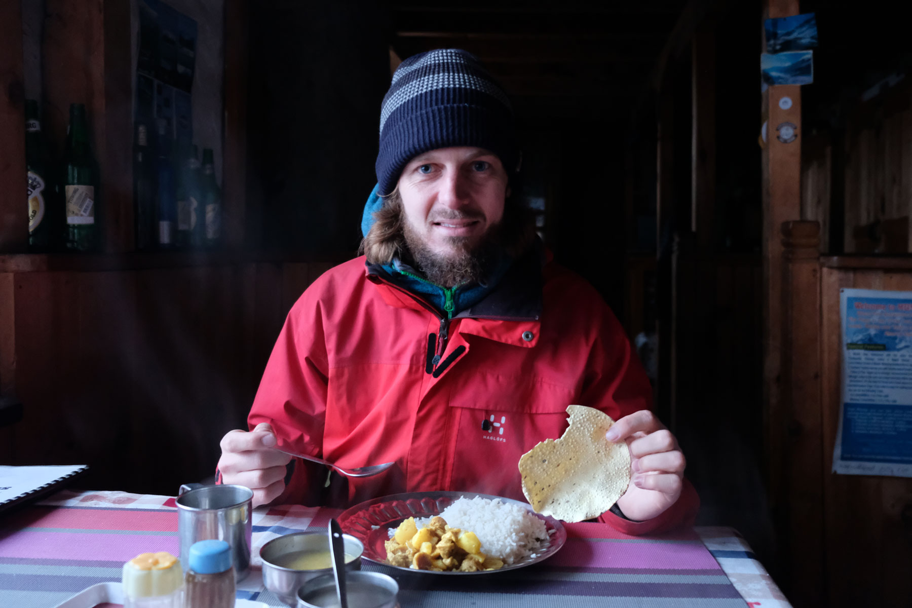 Sebastian sitzt mit Mütze an einem Tisch und isst Dal Bhat.