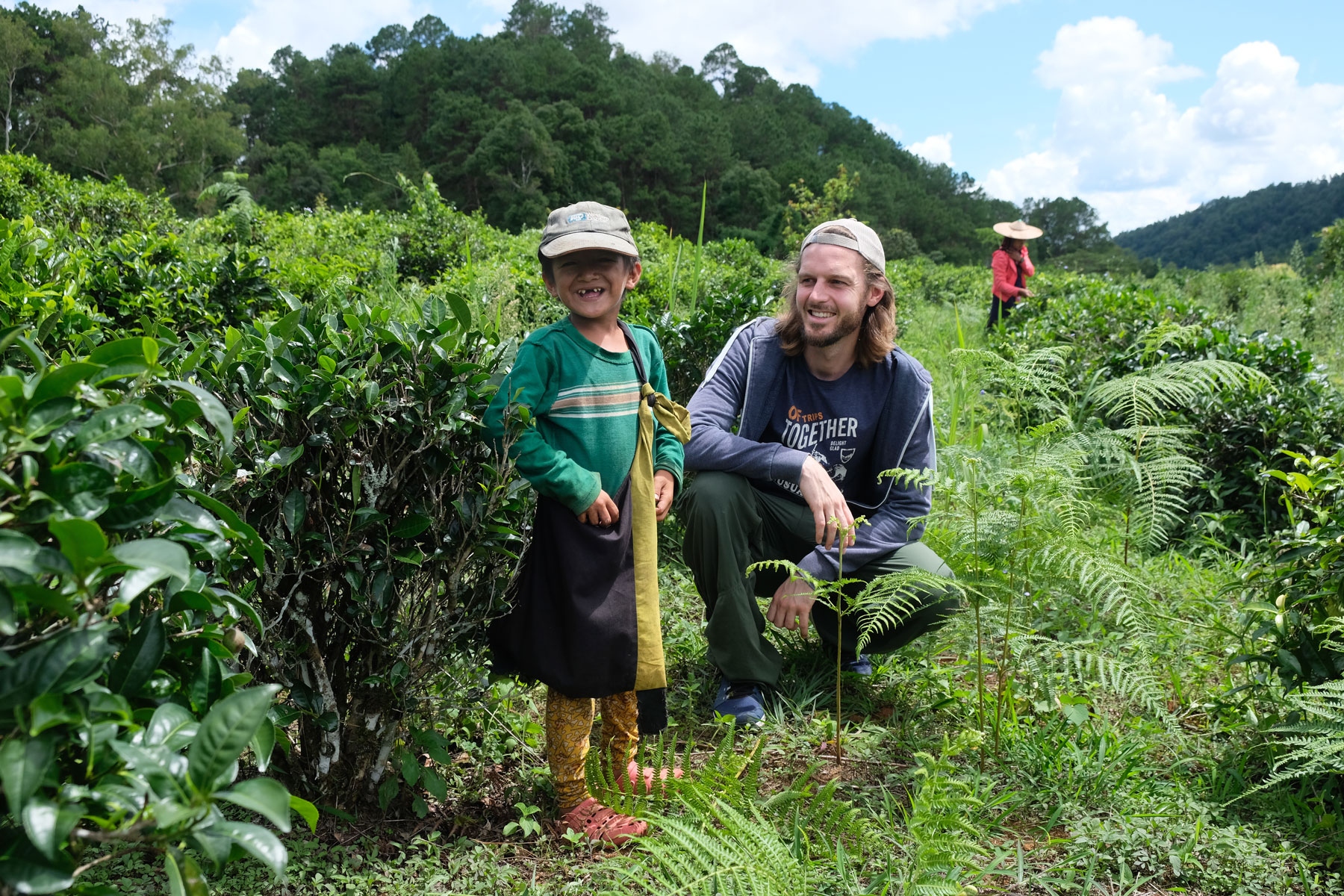Sebastian hockt neben einem lächelnden Jungen inmitten einer Teeplantage in Ban Rak Thai.