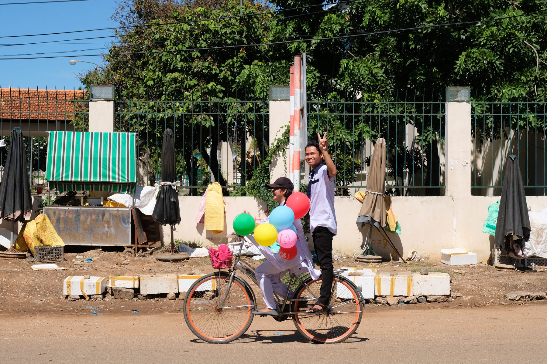 Ein Mädchen fährt auf seinem Fahrrad und hält in der Hand bunte Luftballons. Ein Junge fährt auf dem Fahrrad mit und winkt.