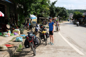 Leo steht mit den beiden Fahrrädern an Straßenständen an der Straße in Vietnam.
