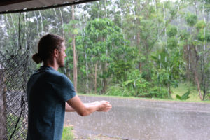 Sebastian steht unter einem Dach und wäscht sich seine Hände am vom Dach strömenden Regenwasser.