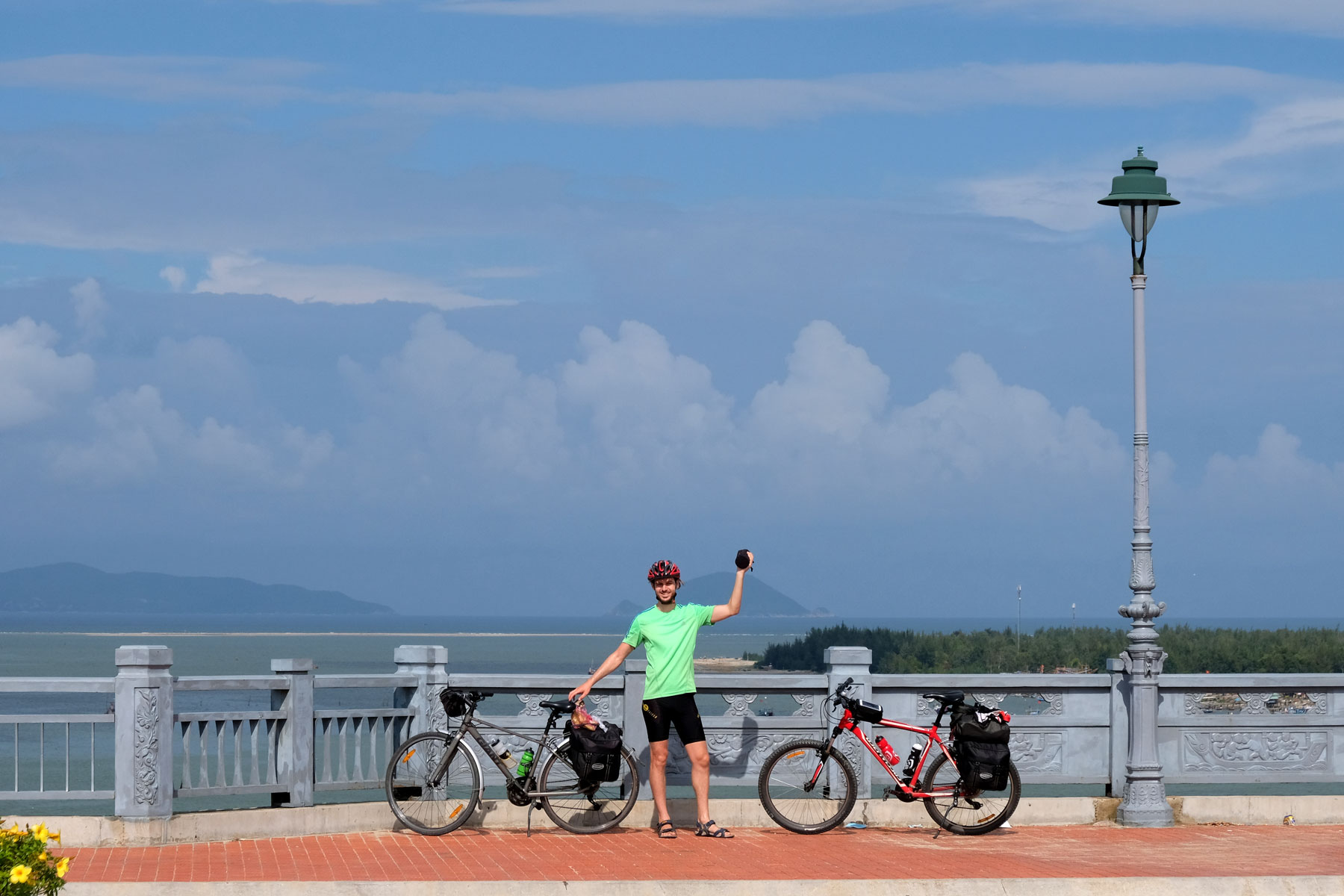 Sebastian steht mit den zwei Fahrrädern auf der großen Brüce vor Hoi An in Vietnam, hinter ihm das Meer.