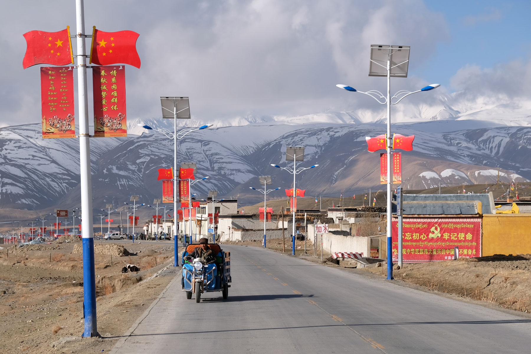 Ein Mann fährt mit seinem elektrischen Dreirad über eine Straße in Tibet, rechts und links von ihm chinesische Flaggen.