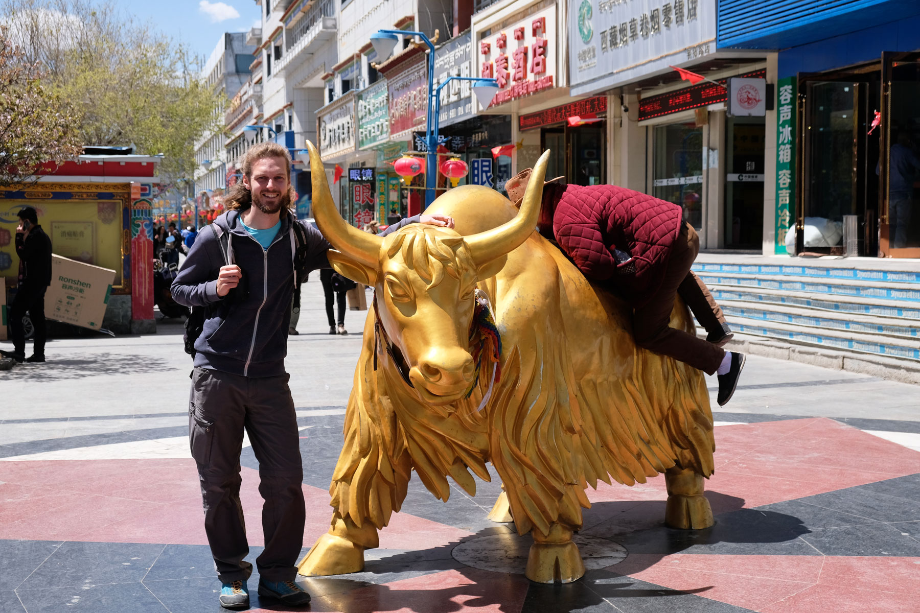 Sebastian steht für ein Foto vor dem goldenen Stier Lhasas, während ein Chinese auf den Stier springt.