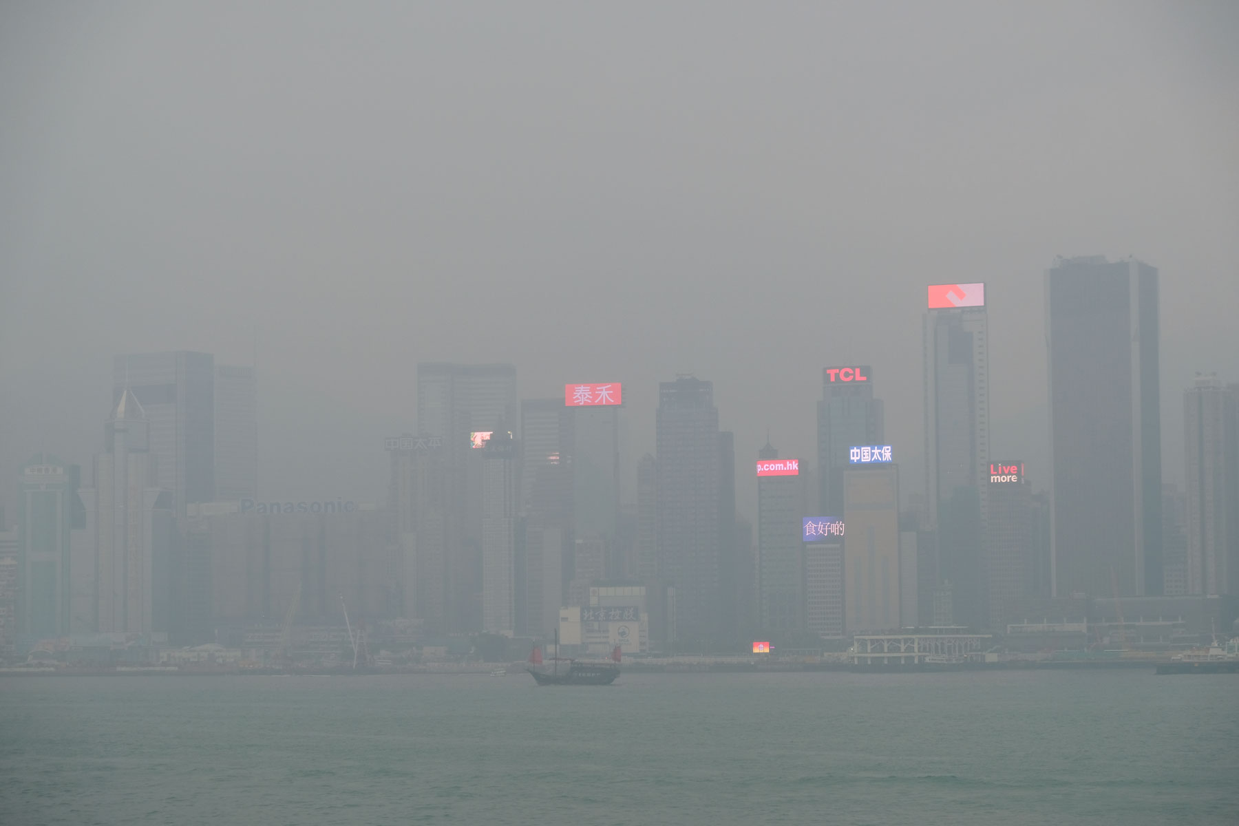 Ein Schiff mit roten Segeln fährt vor der Skyline Hongkongs vorbei, die im Nebel versinkt.