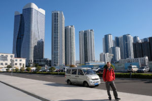 Sebastian steht vor einigen Hochhäusern Qingdaos in China.