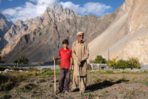 Ein alter Mann und ein Junge stehen auf einem Feld vor einer Bergkulisse im Hunza Tal.